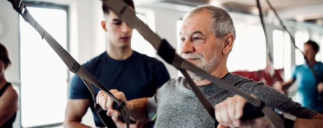 Ein älterer Herr führt eine Übung am Slingtrainer aus und wird von einem jungen Fitnesstrainer betreut