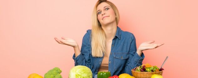 Eine Frau sitzt an einem Tisch auf dem viel Obst und Gemüse liegt und macht mit den Händen eine fragende Bewegung