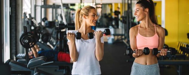 Zwei junge Frauen trainieren mit Kurzhanteln im Fitnessstudio und schauen sich an.